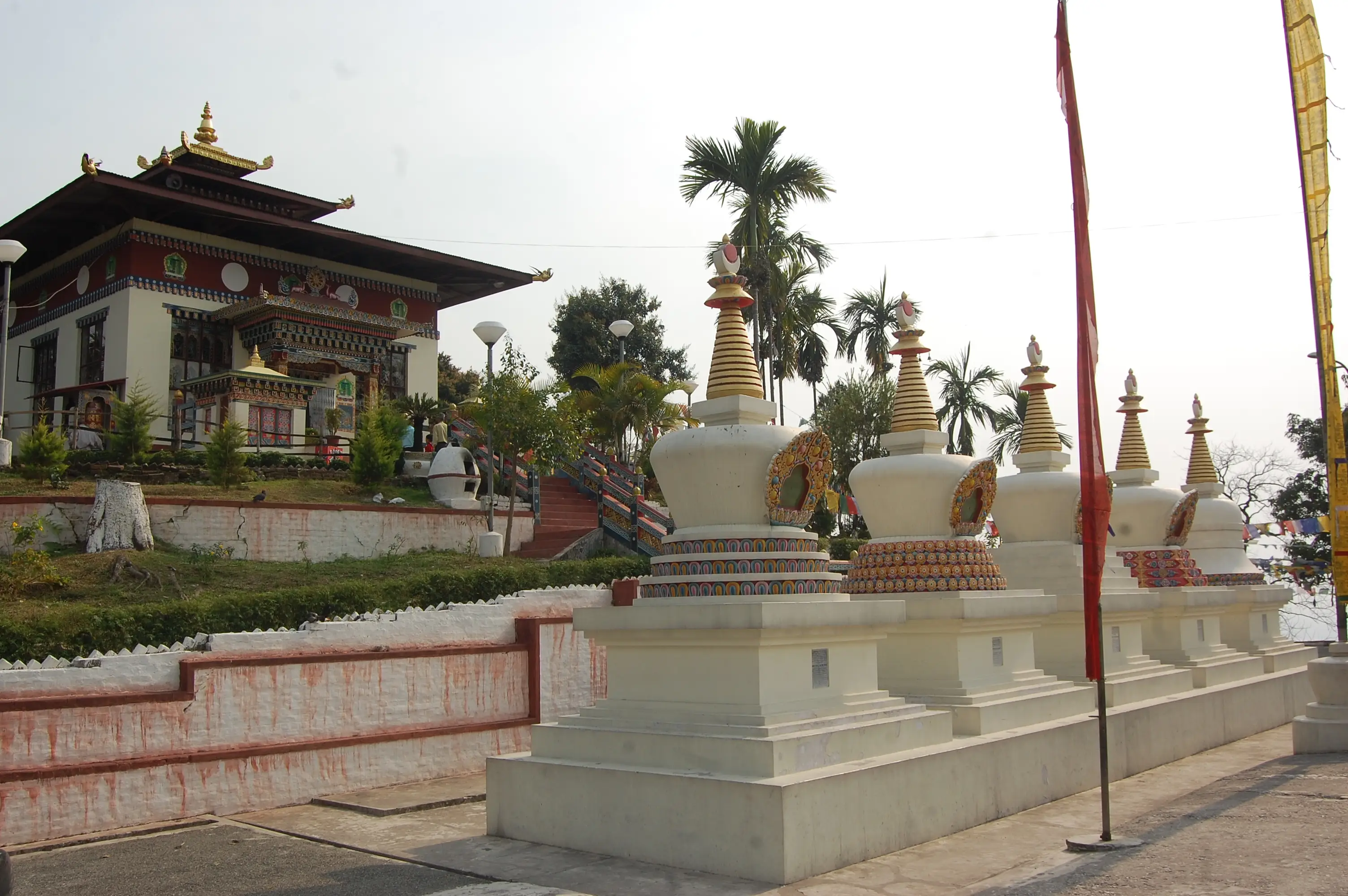 A row of white chortens and the painted temple of Kharbandi Gompa on the hill above Phuentsholing, palm trees behind them in the subtropical haze